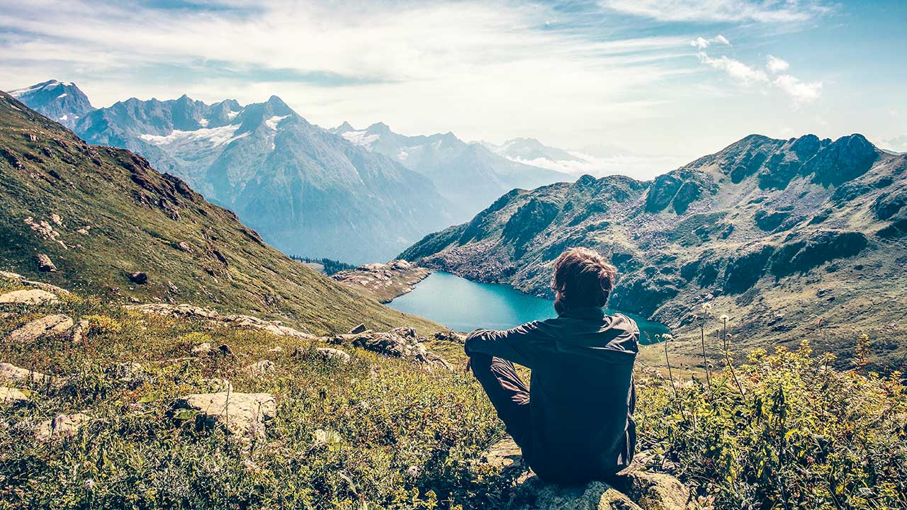 Mann sitzt auf einer Wiese und blickt auf ein Gebirge mit Bergsee
