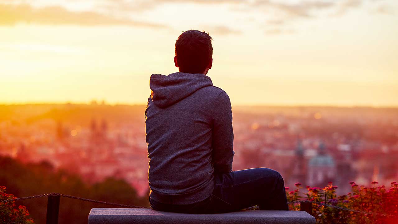 Person sitzt bei Sonnenaufgang auf einem Steinsitz und blickt bei Sonnenuntergang auf die untenliegende Stadt