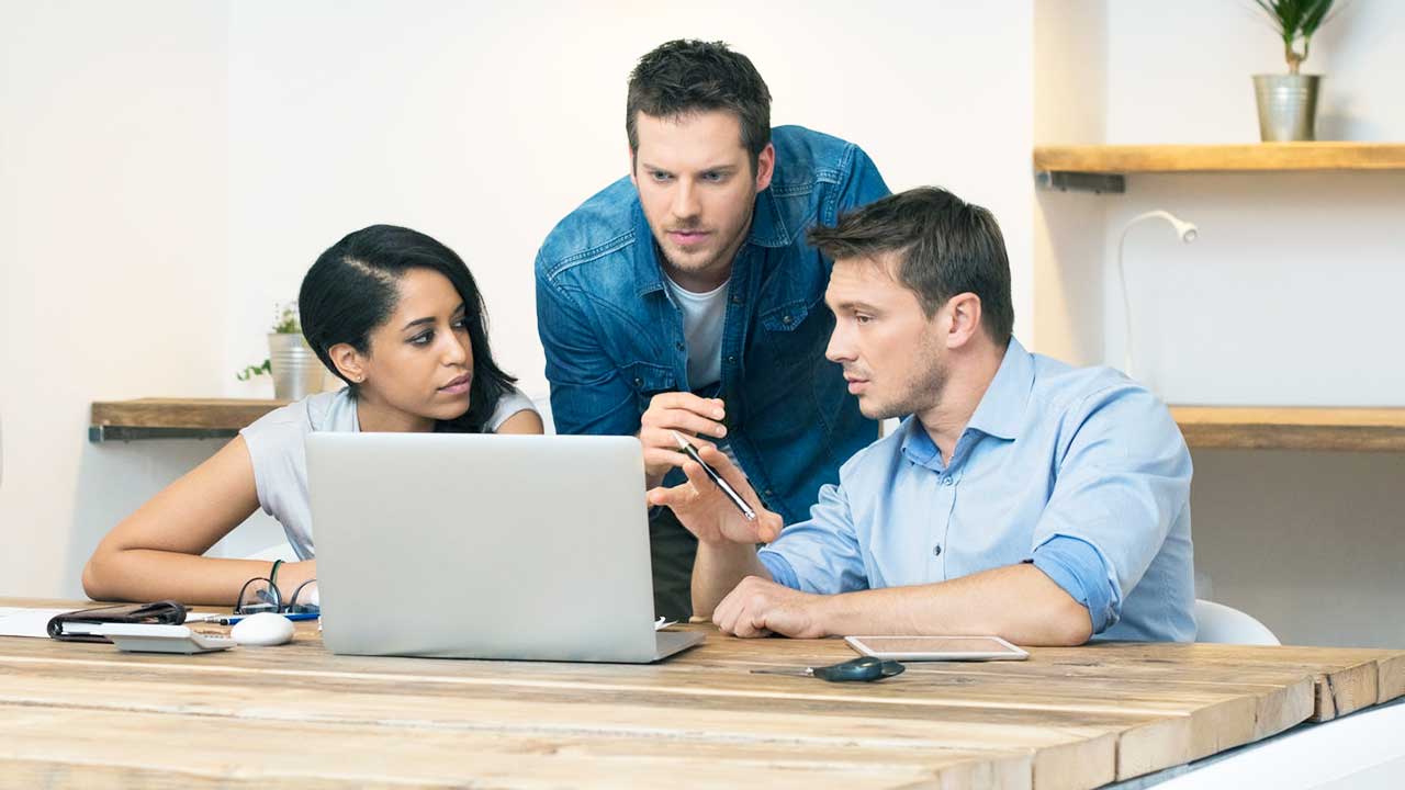 Eine Frau und zwei Männer diskutieren an einem Büroarbeitsplatz mit Computer