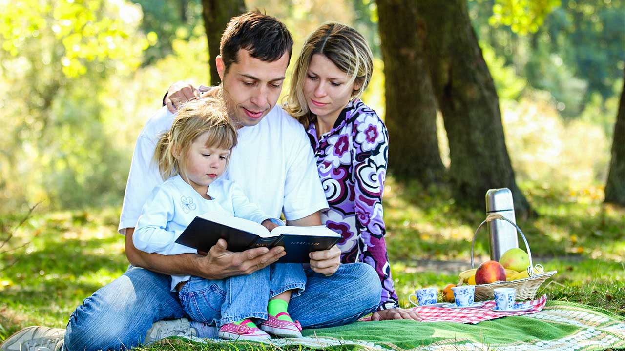 Eltern mit Mädchen sitzen auf einer Picknickdecke und lesen in der Bibel
