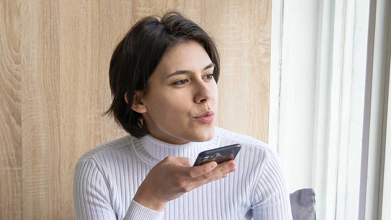 Frau sitzt beim Fenster und nimmt mit ihrem Smartphone gerade eine Sprachnachricht auf