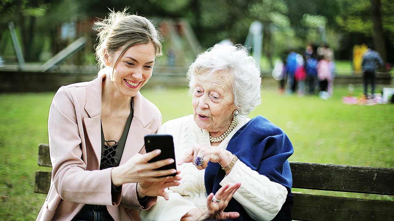 Junge Frau und Seniorin sitzen auf einer Parkbank und schauen gemeinsam etwas auf dem Smartphone an