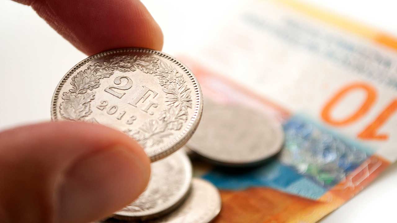 Finger halten Zwei-Franken-Münze, auf einem Tisch liegt weiteres Geld