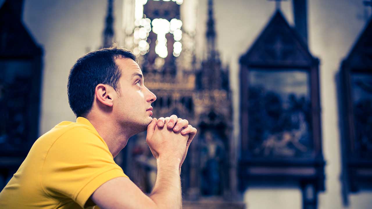 Junger Mann kniet in einer Kirchenbank und richtet seinen Blick nach oben