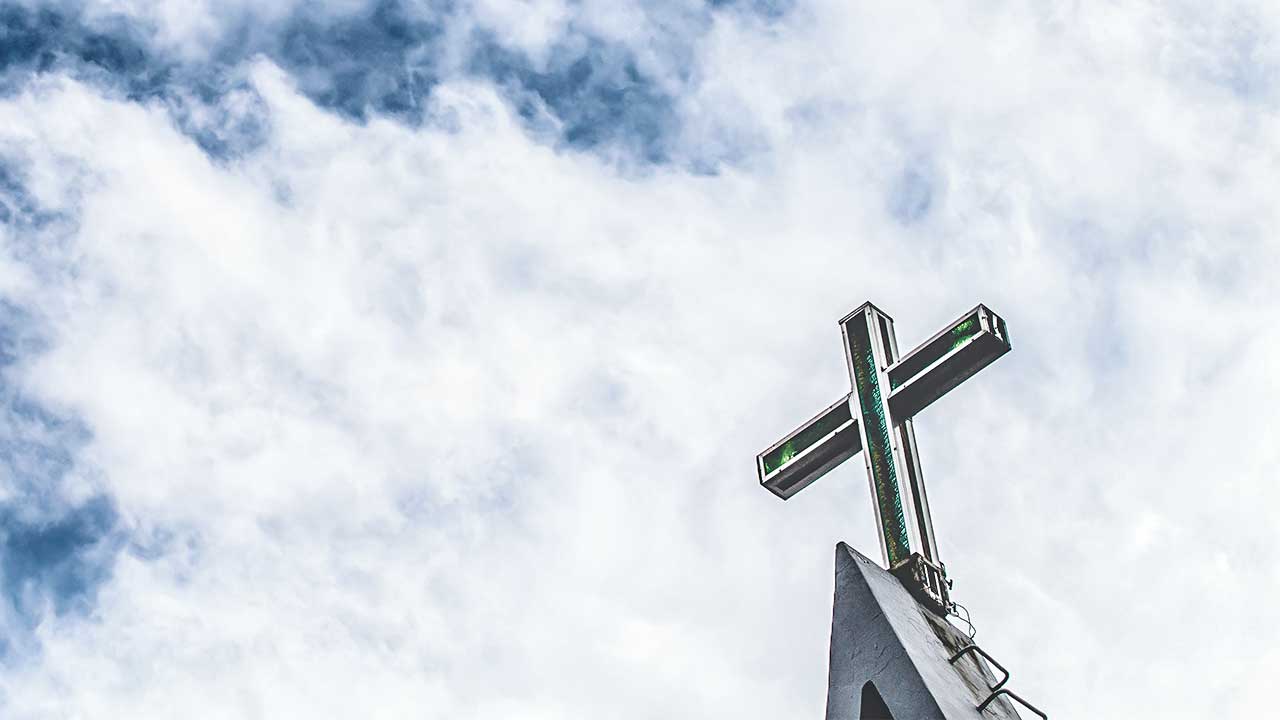 Kreuz auf einem Kirchendach, darüber Himmel mit Wolken
