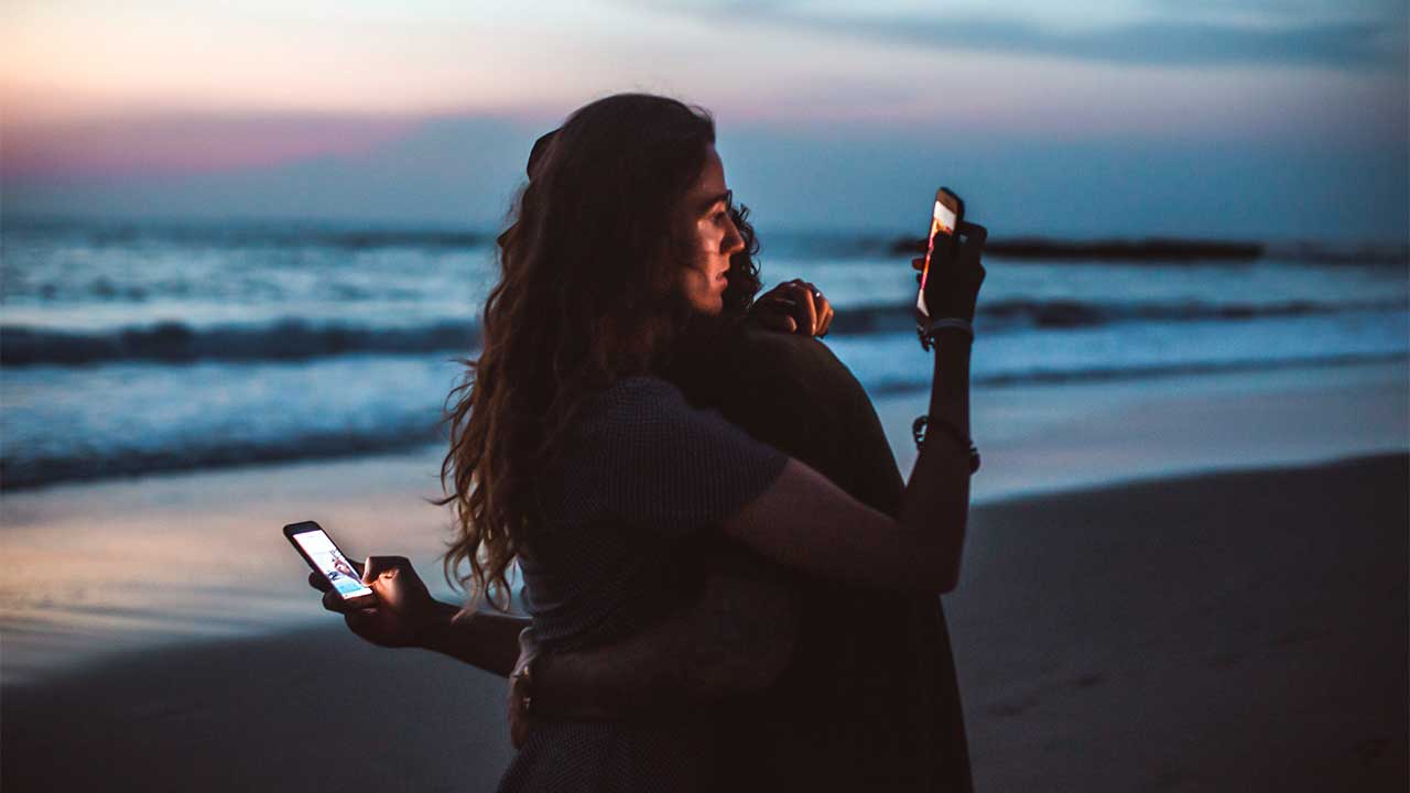 ein Paar umarmt sich an einem Strand und blickt gleichzeitig auf Smartphones